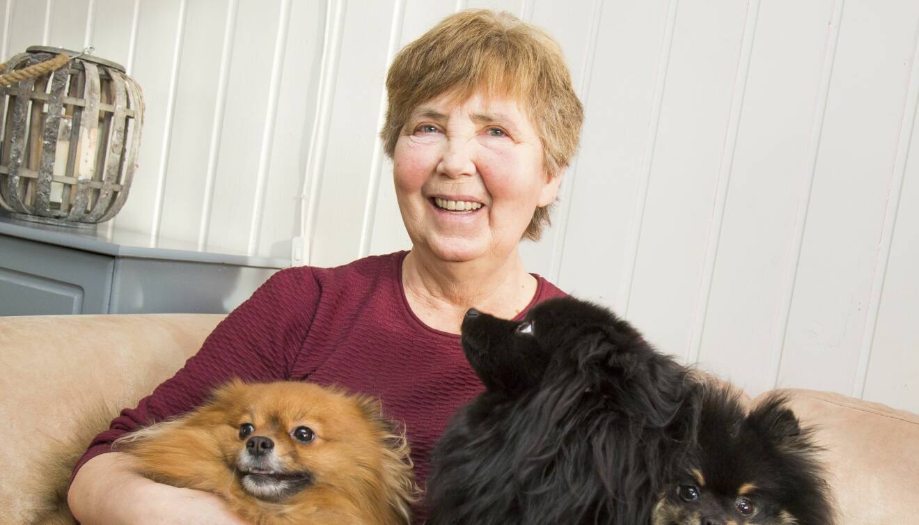 En äldre leende kvinna sitter i en soffa med sina tre hundar i knät.