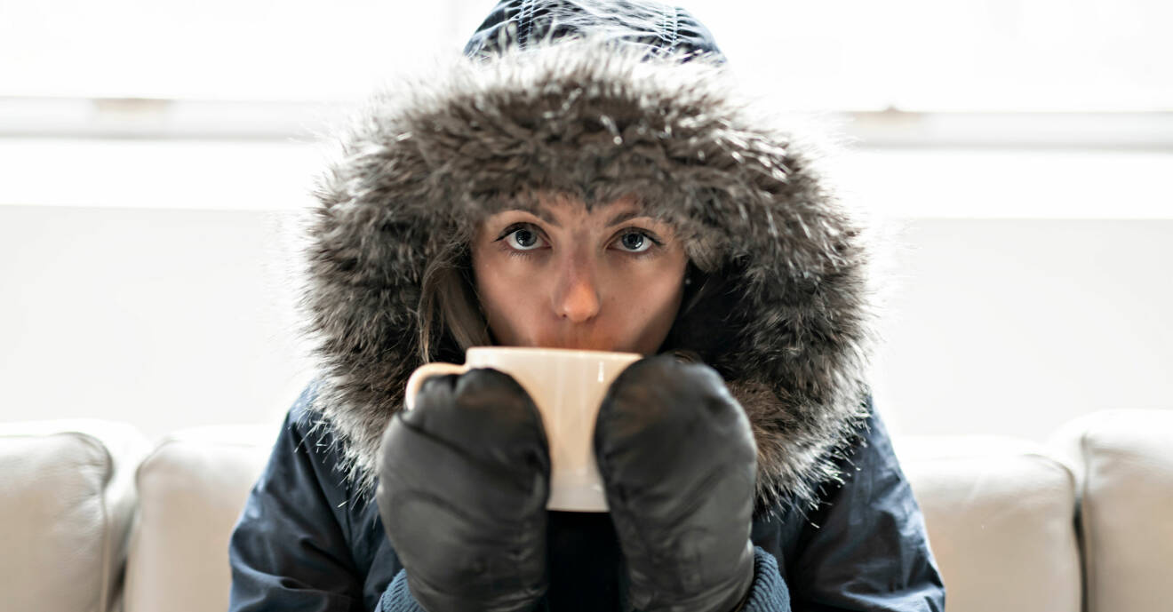 Frusen kvinna i vinterjacka dricker kaffe i soffan.