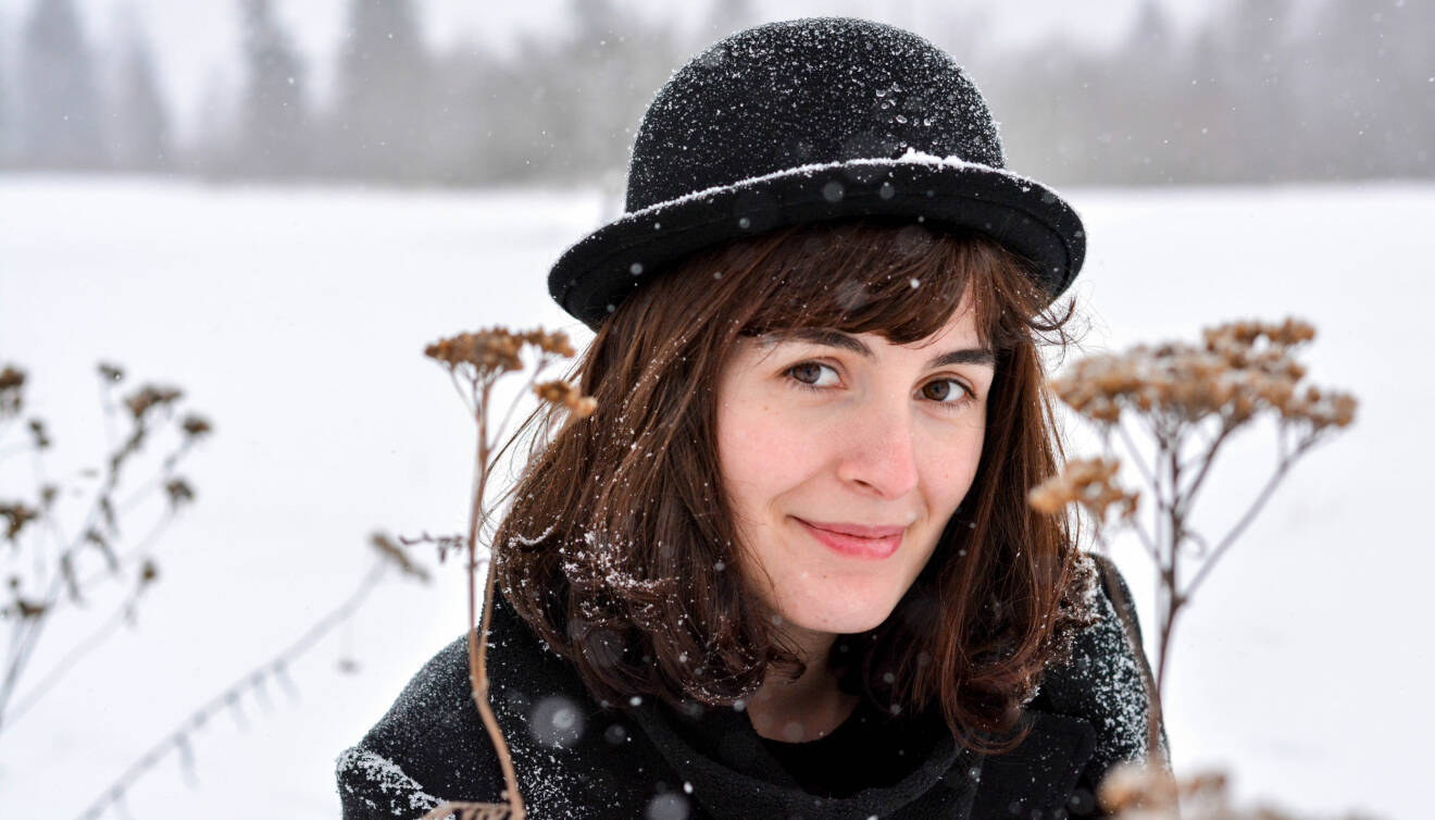 Kvinna i hatt ler mot kameran. I bakgrunden syns snöfall.