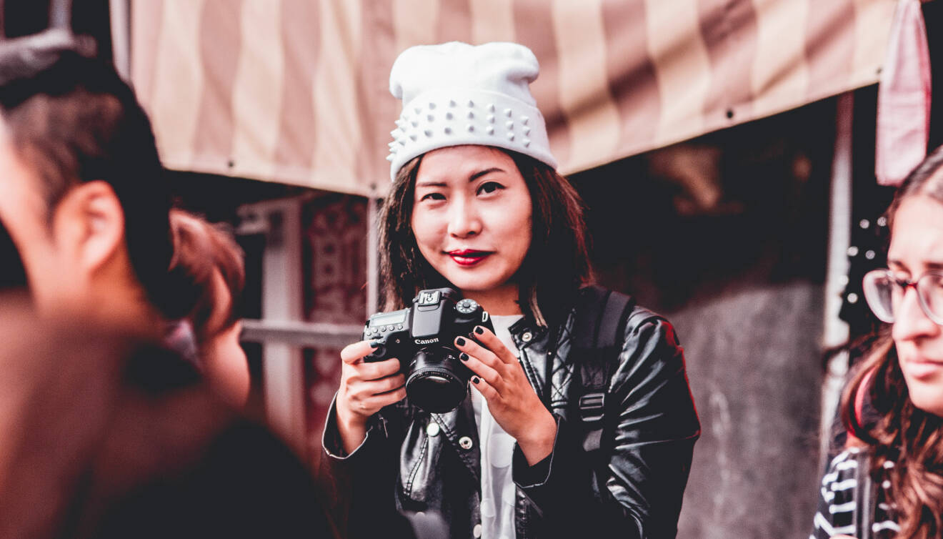 Kvinna håller en kamera i handen och tittar glatt mot fotografen.