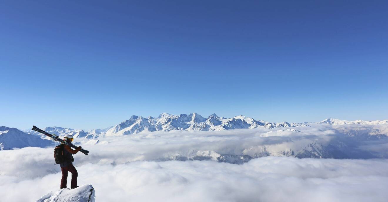 Johanna står längst ut på en klippa med skidor över axeln och blickar ut över alptoppar.