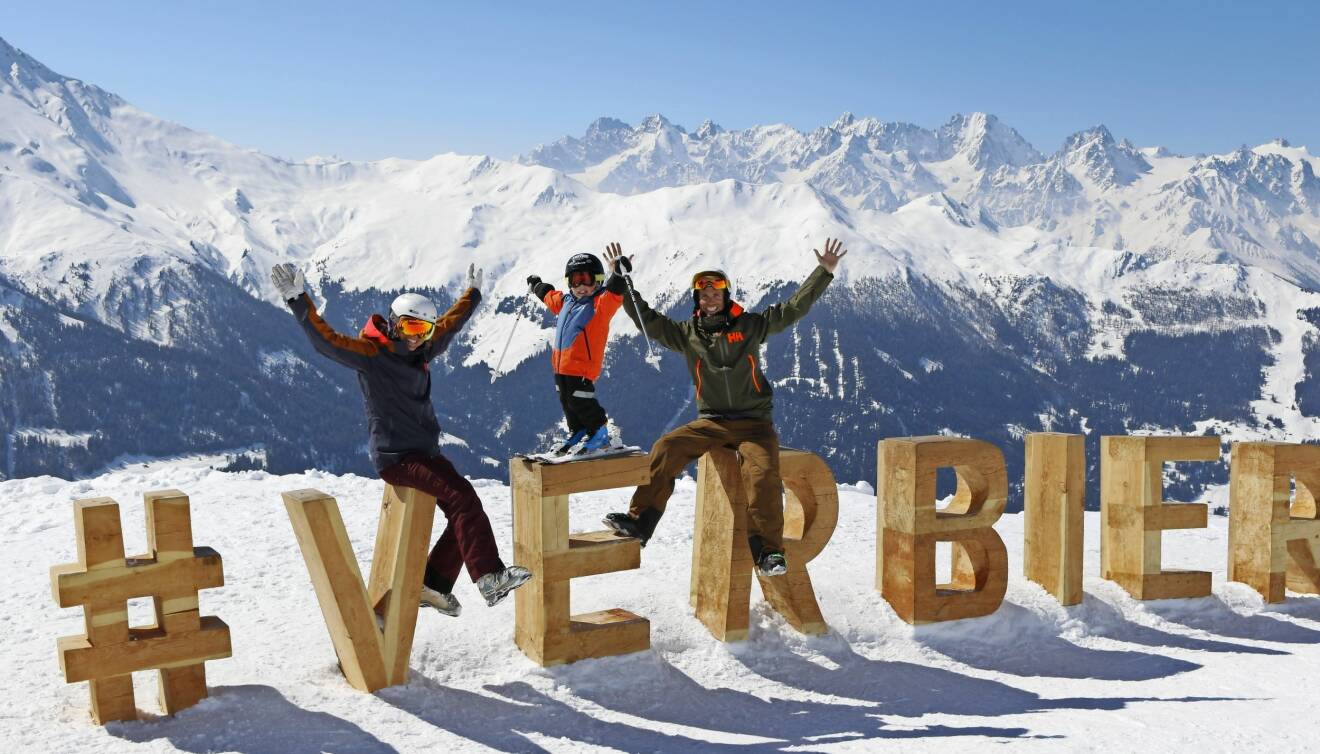 Johanna, Anders och Walter sitter på stora träbokstäver som formar namnet Verbier i en skidbacke.