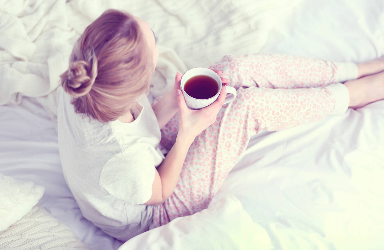 Kvinna sitter i en säng med en kopp te i handen.