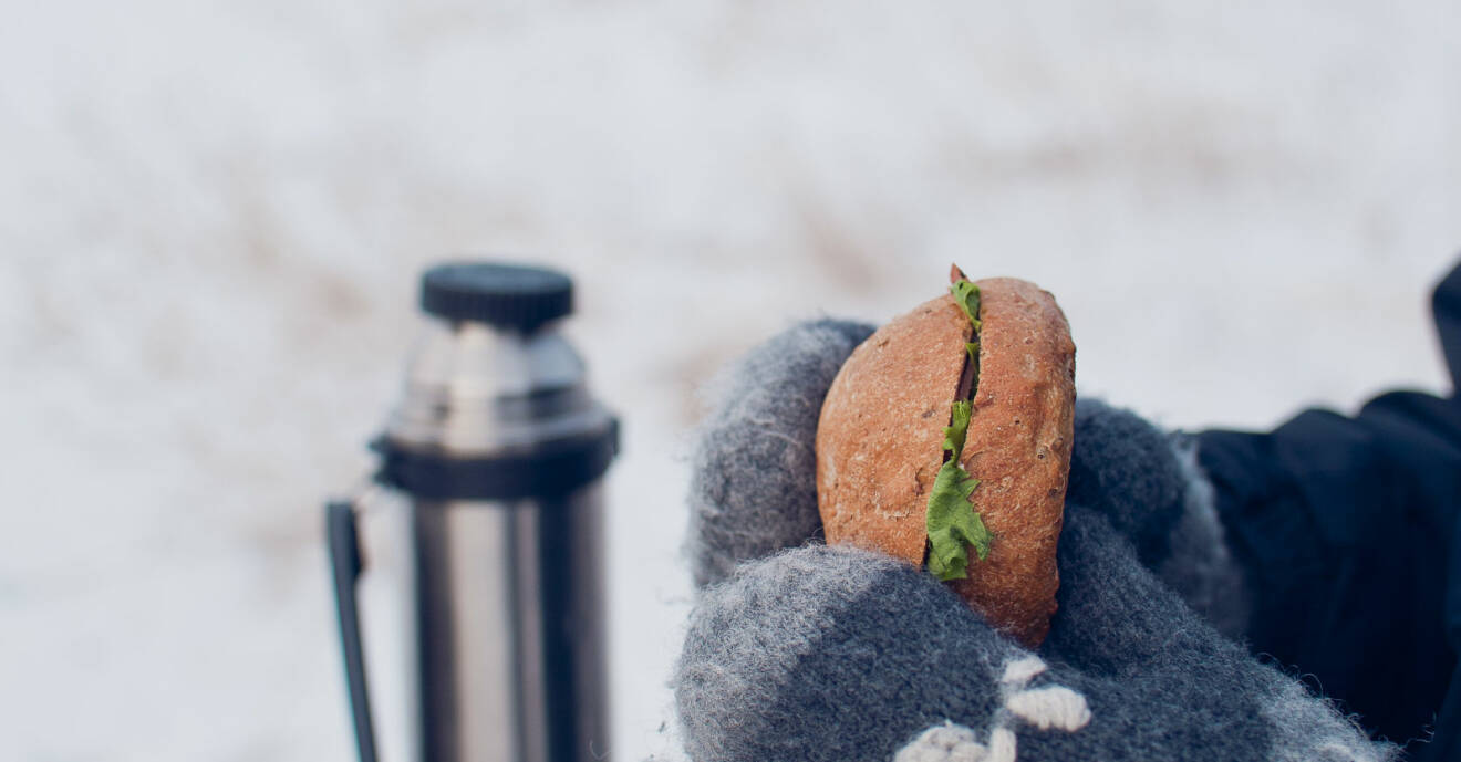 Två händer med vantar som håller i en smörgås utomhus