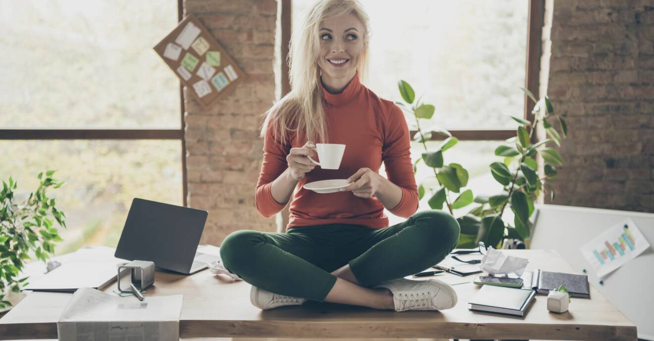 En kvinna sitter i skräddarsits och dricker en kopp te eller kaffe på sitt skrivbord.