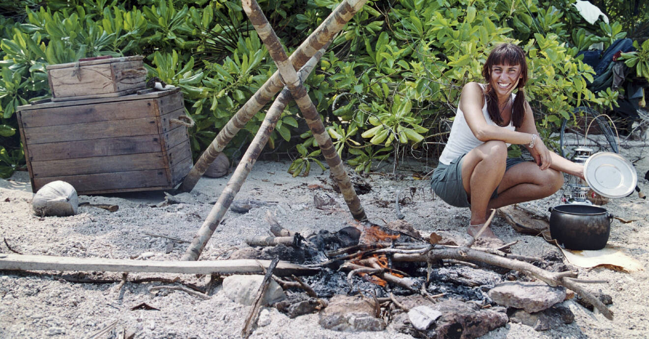 Zübeyde sitter på huk på en strand, bredvid henne finns en öppen eld och hon håller ett lock som hon tagit av från en kastrull