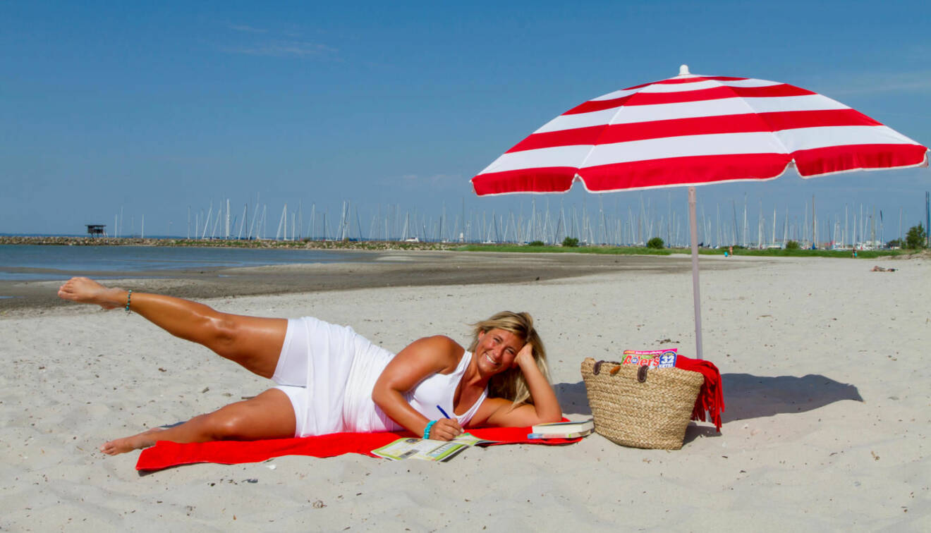 Kvinna ligger på stranden, på en handduk och löser korsord medan hon tränar genom att lyfta benen i sidled.