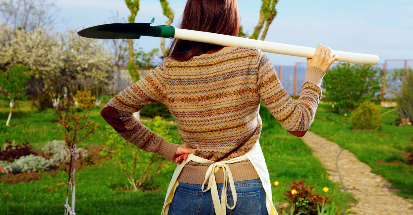En kvinna står med en spade över axlarna och tittar ut över sin trädgård.