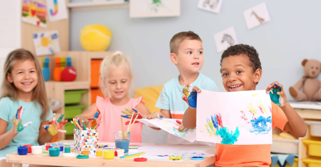 Glada barn målar med fingerfärg vid ett bord på en förskola.