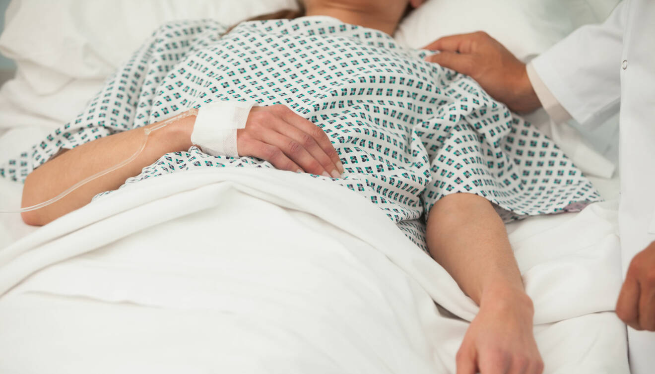 En person ligger i en sjukhussäng med dropp och en läkare håller handen på hennes axel.