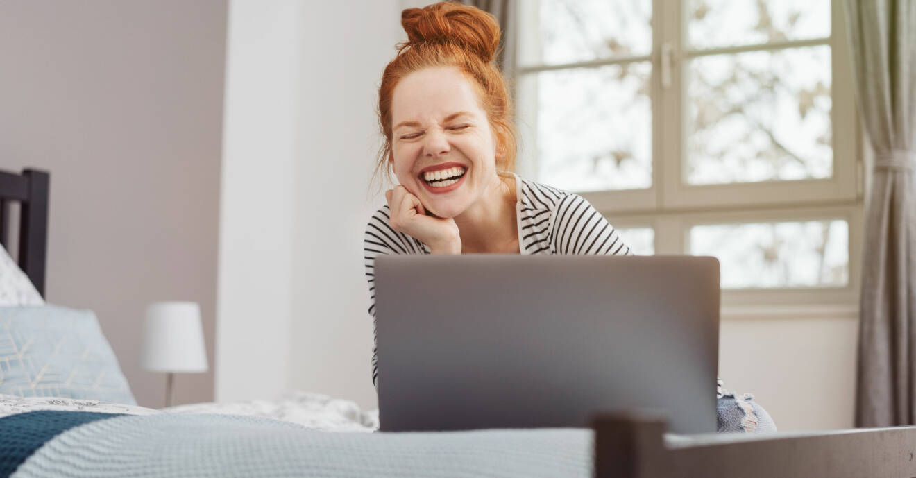 Kvinna sitter på sin säng med datorn uppslagen och skrattar.