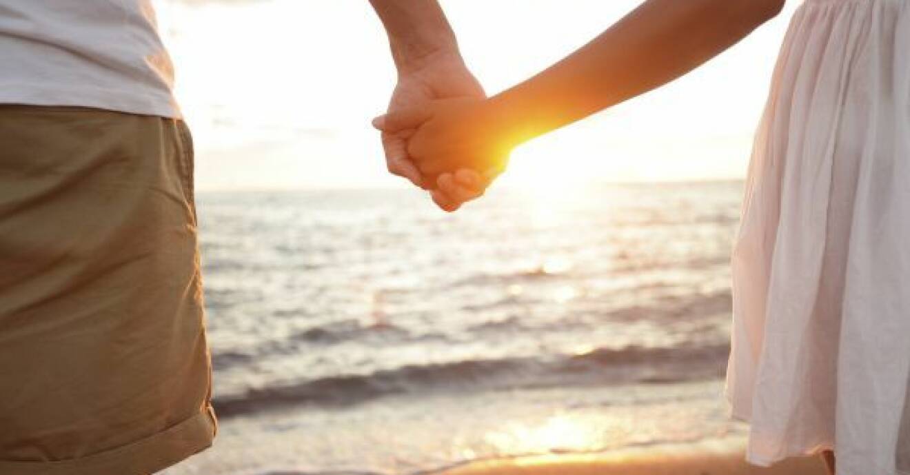 Ett par håller varandra i händerna på en strand, i solnedgången.