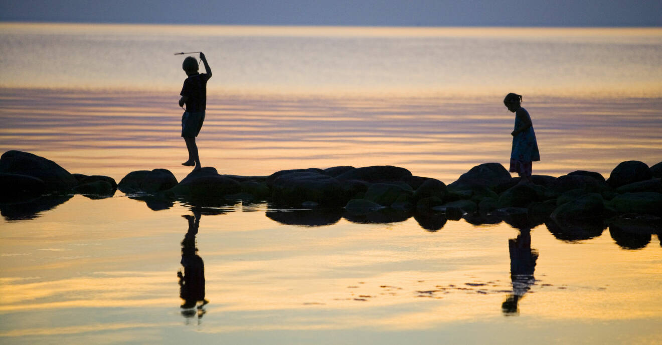 Ett par barn leker på stenar i ett grunt vatten. Det är skymning och de speglas i det stilla vattnet.