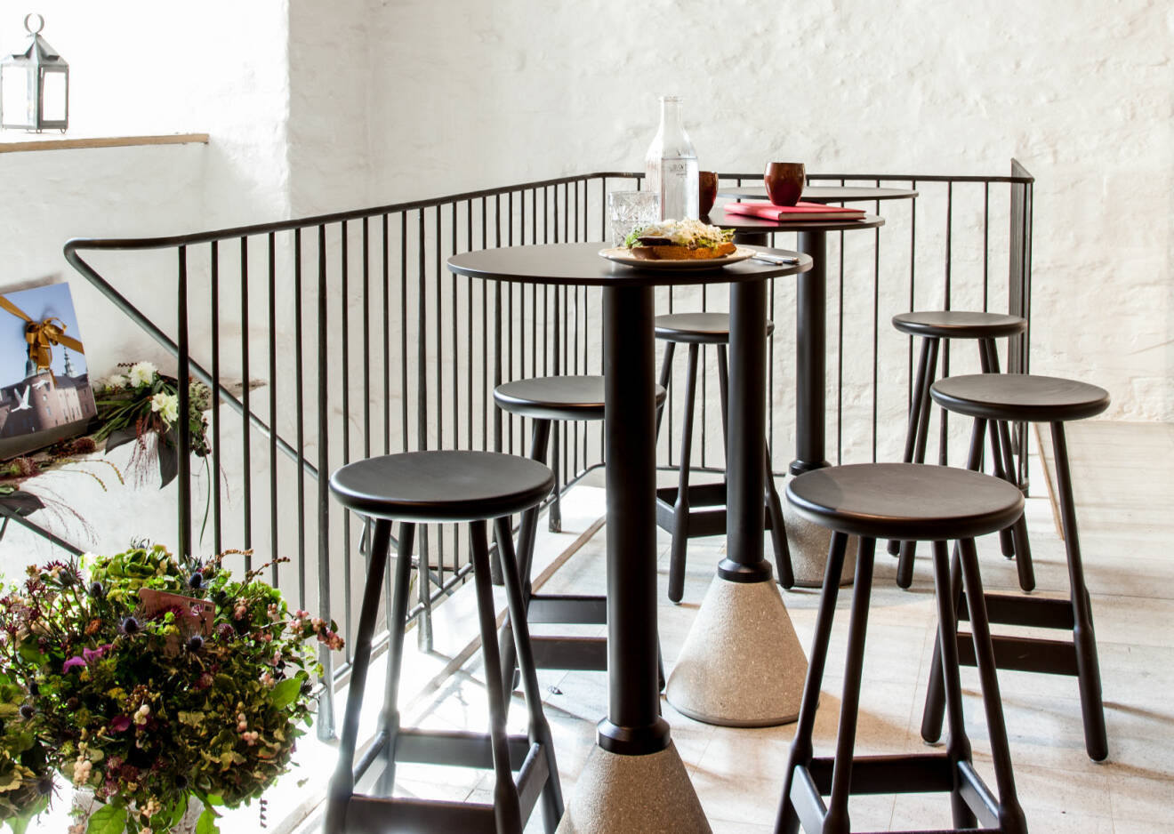Vita stenväggar, mörka stolar och pallar och ett trappräcke i gjutjärn. På borden står en macka och kaffe. I hörnet skytaren stor bukett blommor.