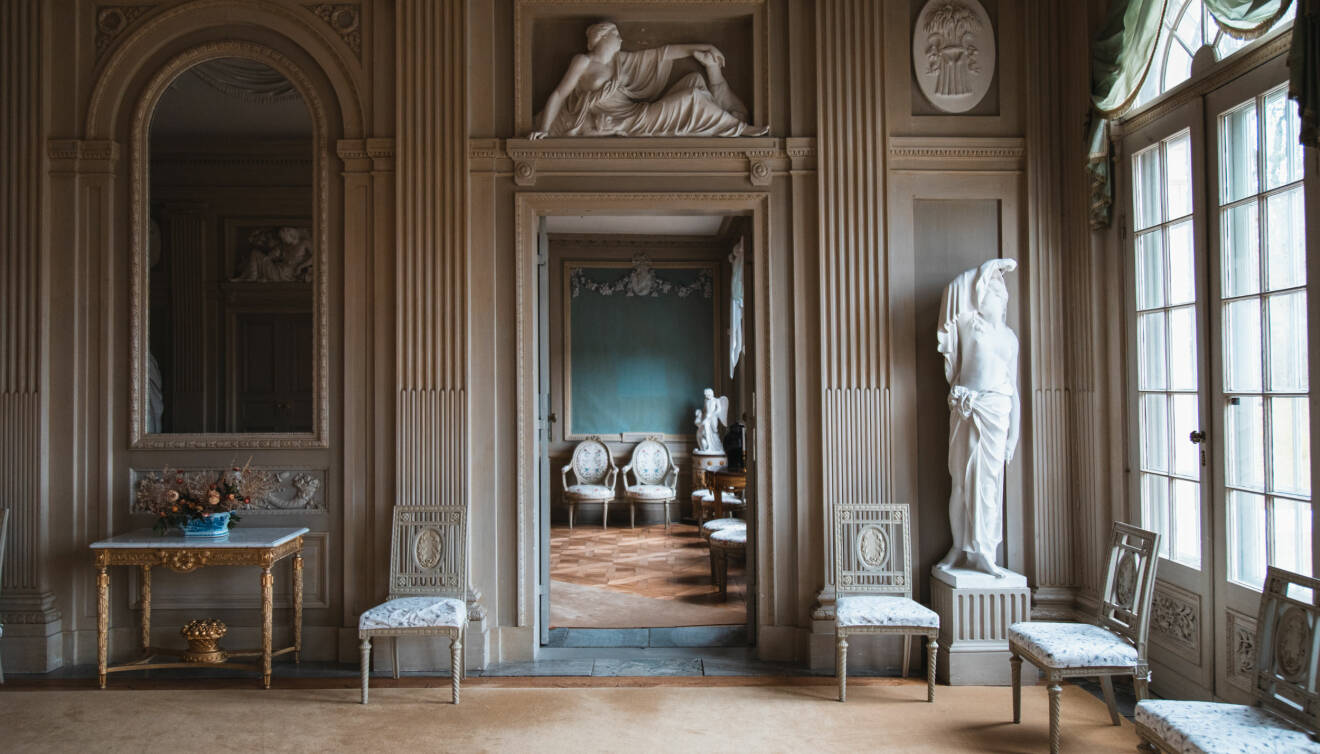 Ett vitt slottsrum med marmorstatyer, ett sirligt avlastningsborg i guld och flera snidade stolar.