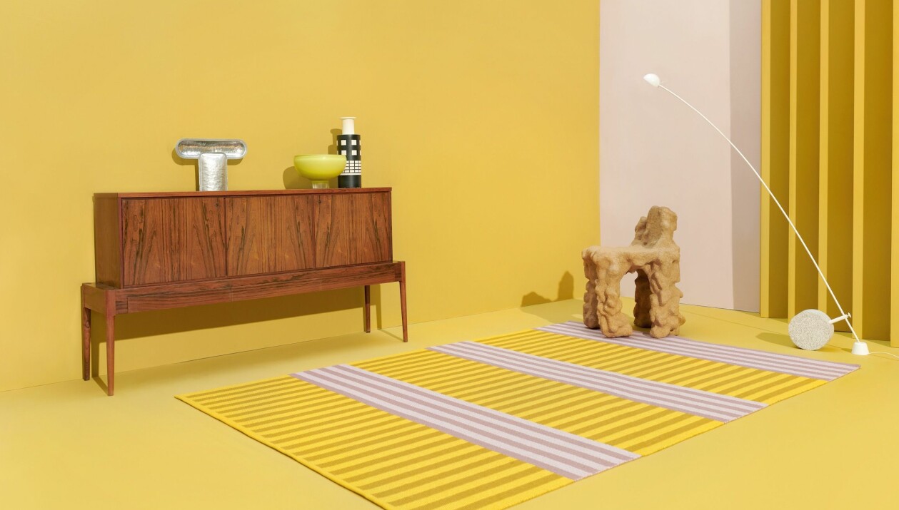I Ett mycket gult rum står en skänk i trä med några prydnadsföremål. På golvet ligger en randig matta i gult och lila.