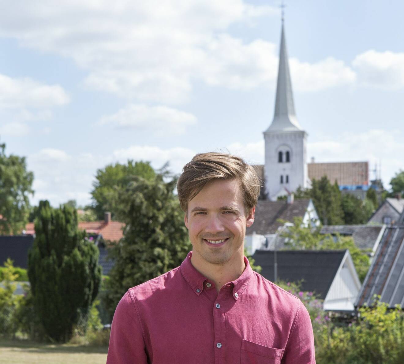Nils Holmqvist i vinröd skjorta och leende framför Anderslövs vitkalkade kyrka.