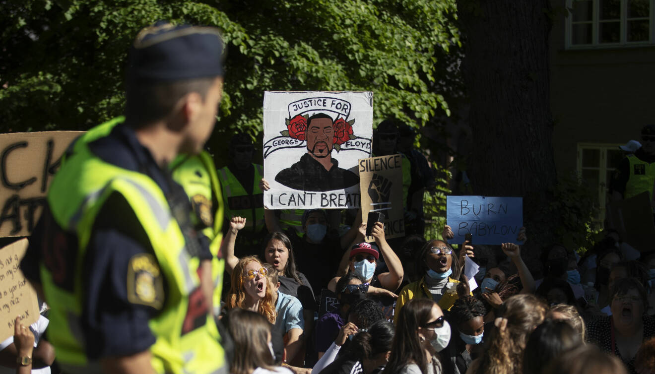 I förgrunden står en polis och tittar bak på en grupp demonstranter som bland annat håller upp en skylt med ett porträtt av George Floyd med texten "Justice for George" och "I can't breathe".