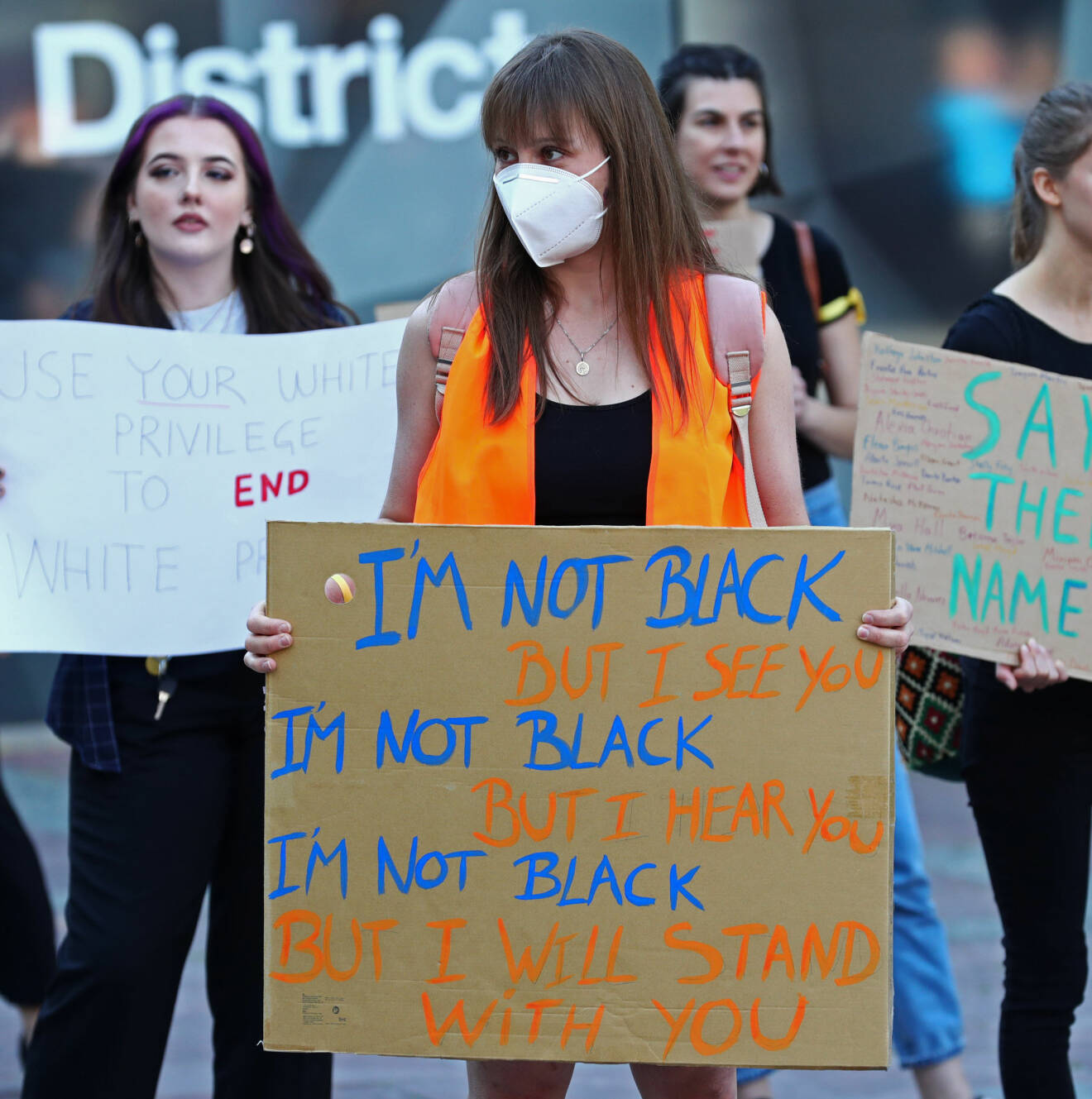 En kvinna med ansiktsmask håller upp en skylt med texten "I am not black, but I see you. I am not black, but I hear you. I am not black, but I will stand with you".