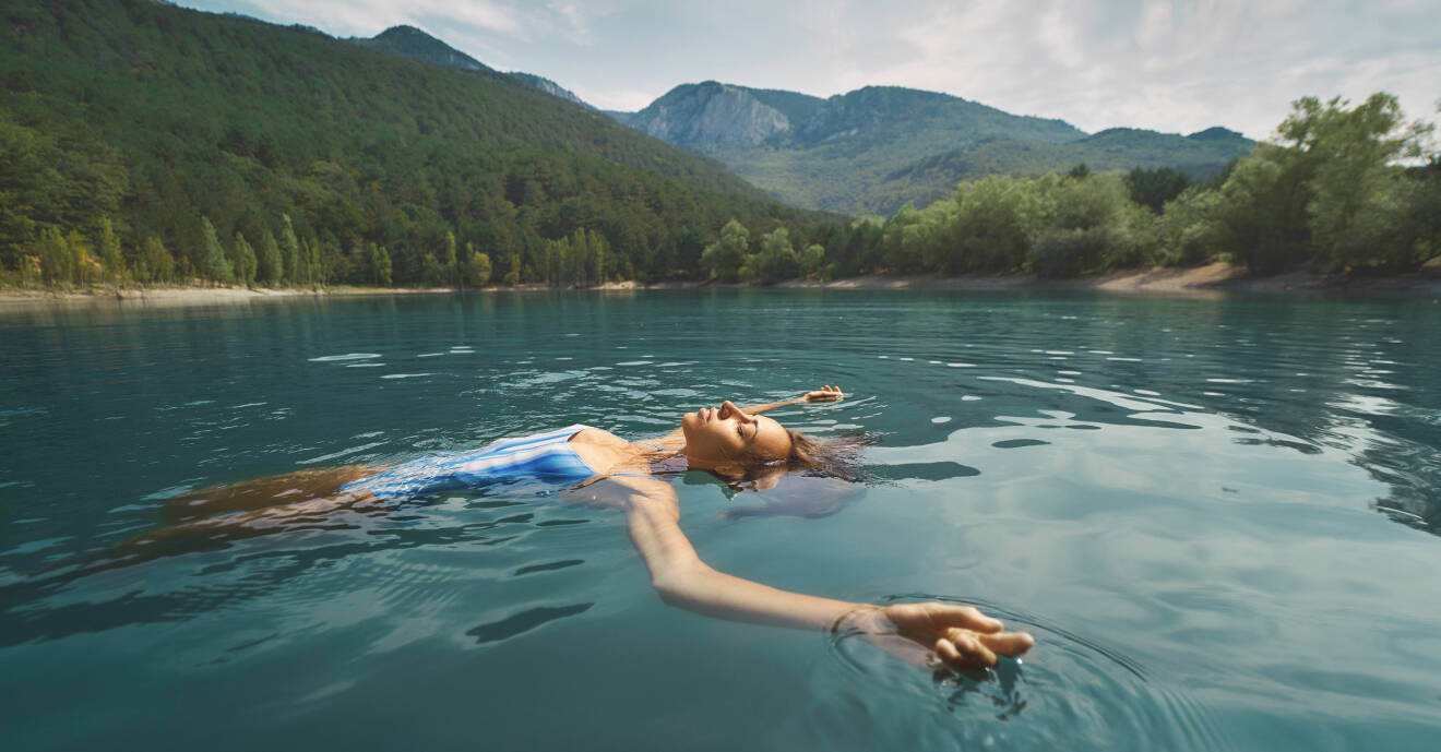 En kvinna flyter avslappnat på rygg i en skogssjö med blått vatten.