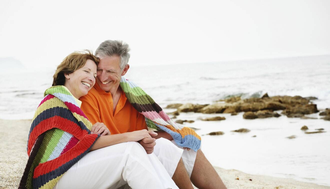 Ett medelålders par sitter på en strand nära varandra under en filt.