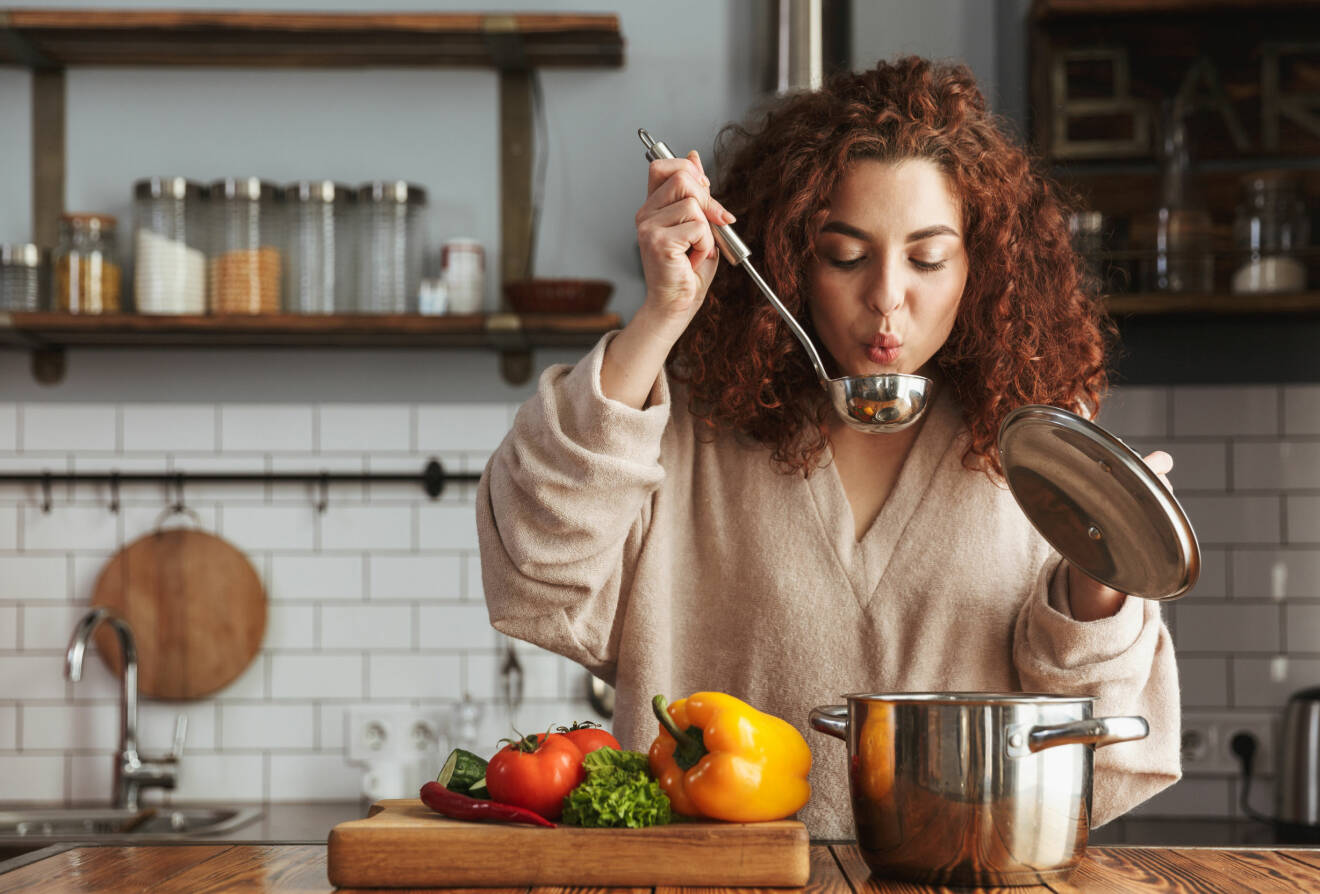 En rödhårig kvinna står i ett kök och provsmakar maten direkt ur soppsleven.