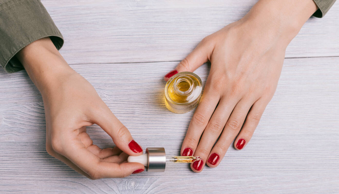 Ett par händer med rött nagellack droppar olja på nagelbanden.