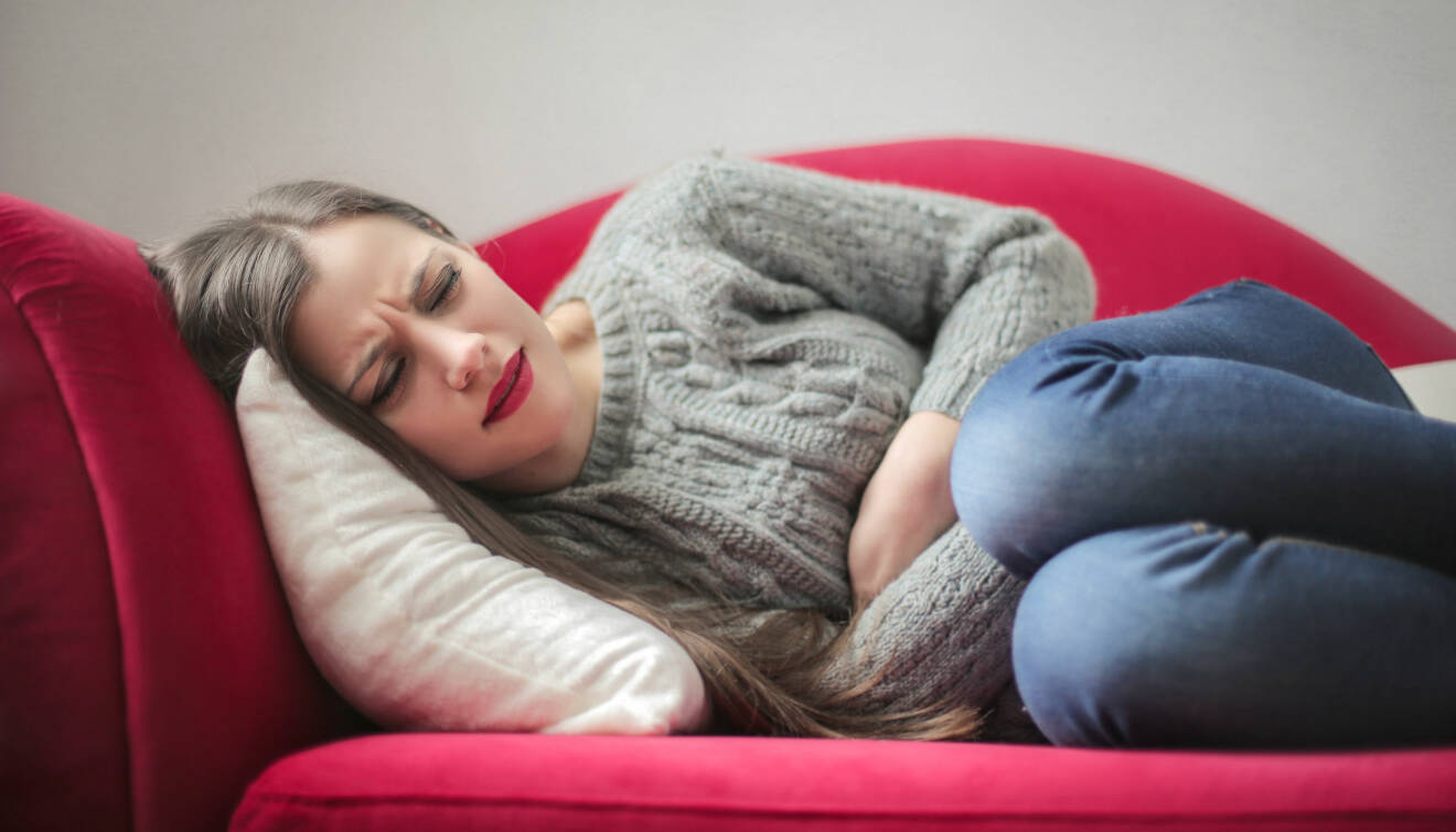 Kvinna ligger på röd soffa och tar sig för magen av smärta.