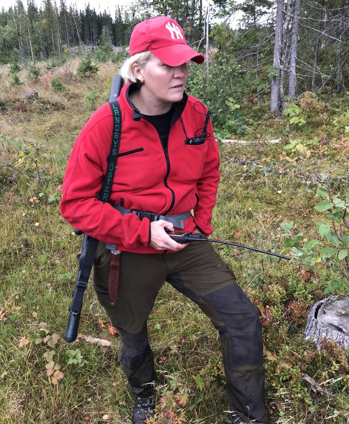 En kvinna står med gevär och walkie talkie i skogen.