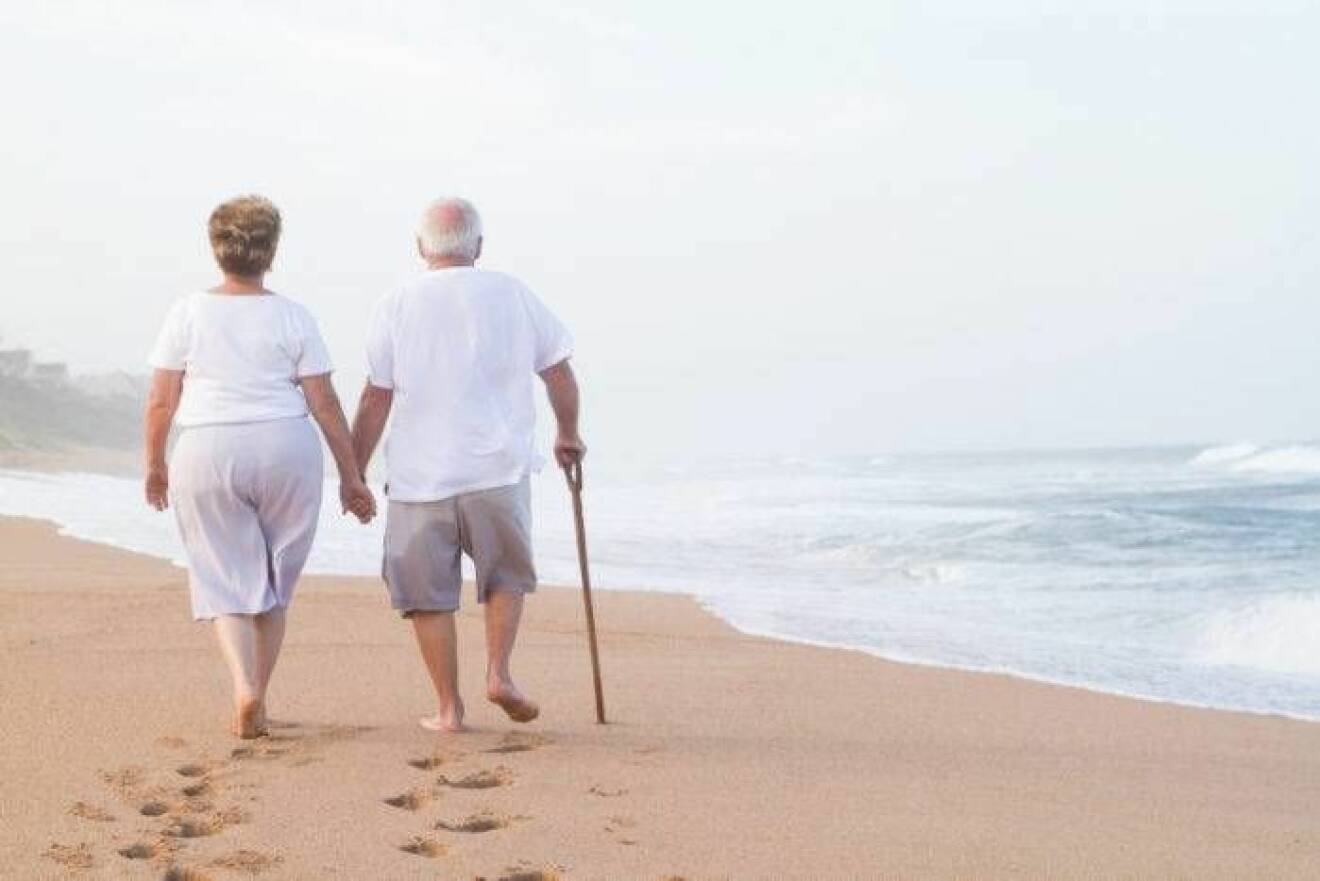 Äldre par går hand i hand på strand.
