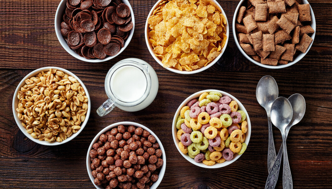 Frukostflingor och müsli kan innehålla rikliga mängder socker. Sex skålar med olika frukostflingor, tre skedar och en kanna mjölk på ett träbord fotograferade rakt uppifrån.