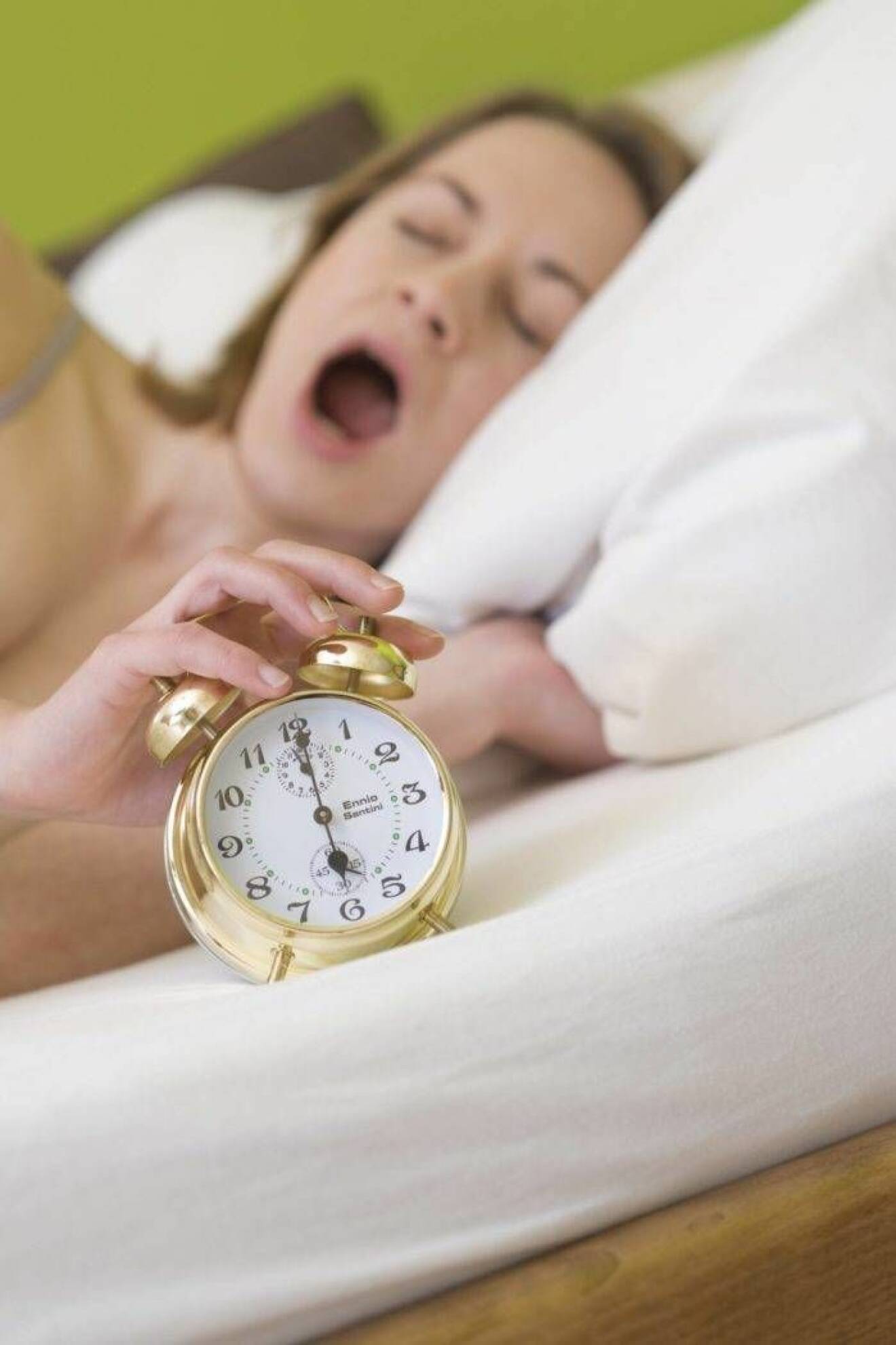 En kvinna som gäspar i sängen bredvid sin väckarklocka