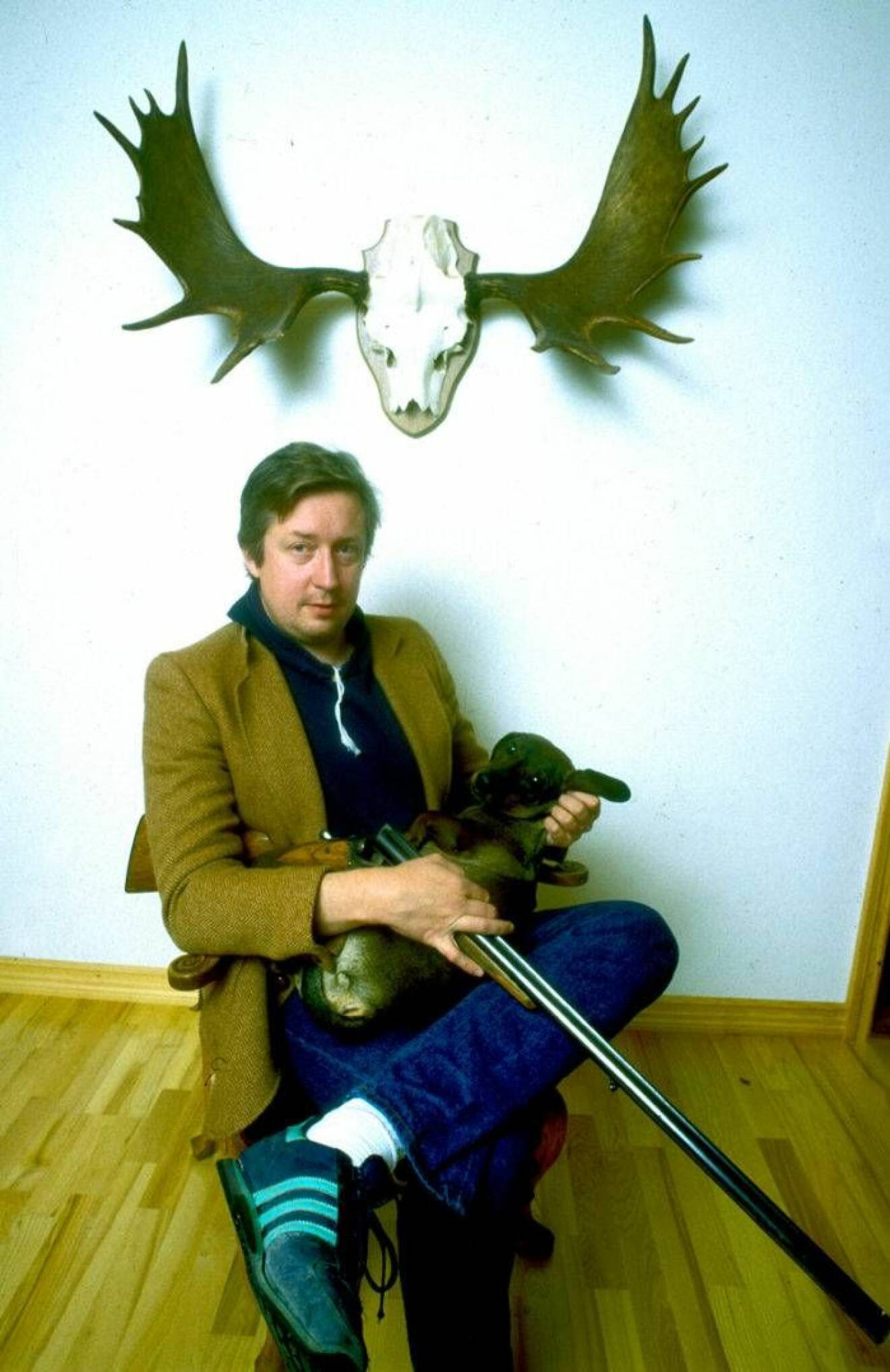 Leif GW Persson sittandes på stol inomhus med jaktgevär i knäet.