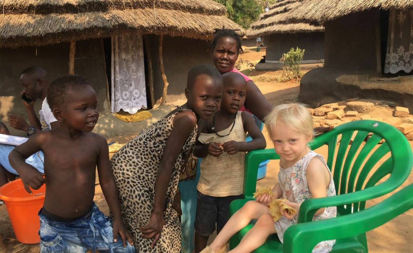 Fyra barn och en kvinna i en by i Uganda med hyddor i bakgrunden.