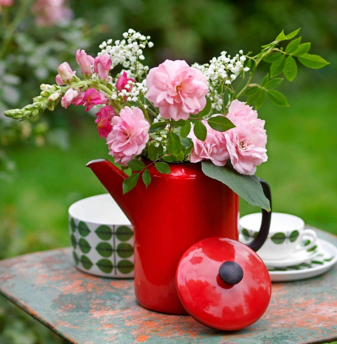 Emaljerad röd kanna med blommor står på ett bord tillsammans med delar ur Stig Lindbergs servis Berså. 