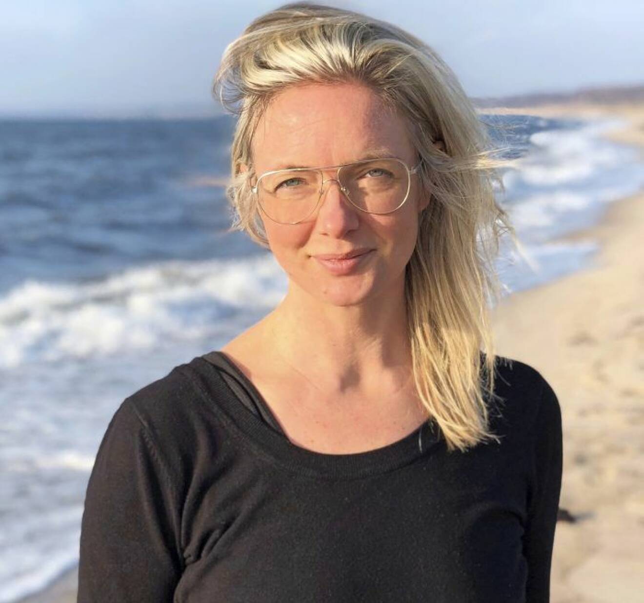 Malin Skogström, blond med långt hår och glasögon i svart tröja vid strand med skummande vågor.