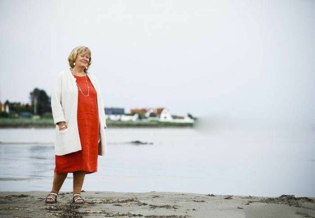 Medelålders kvinna i blond page, med röd klänning och vit kofta står vid havsvik.