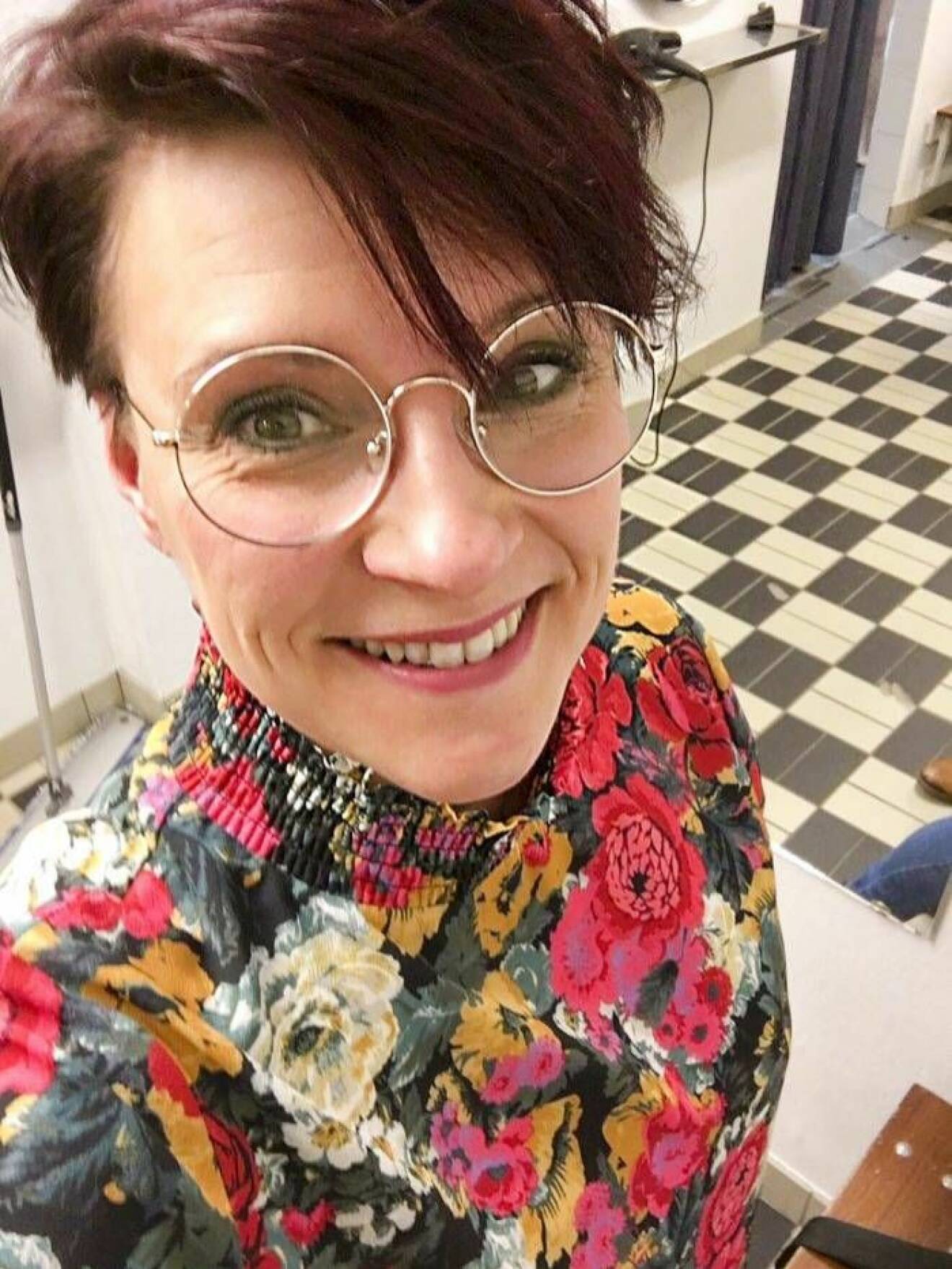 Selfie av glad och pigg kvinna med stormönstrad topp, runda glasögon och kort mörkt hår.