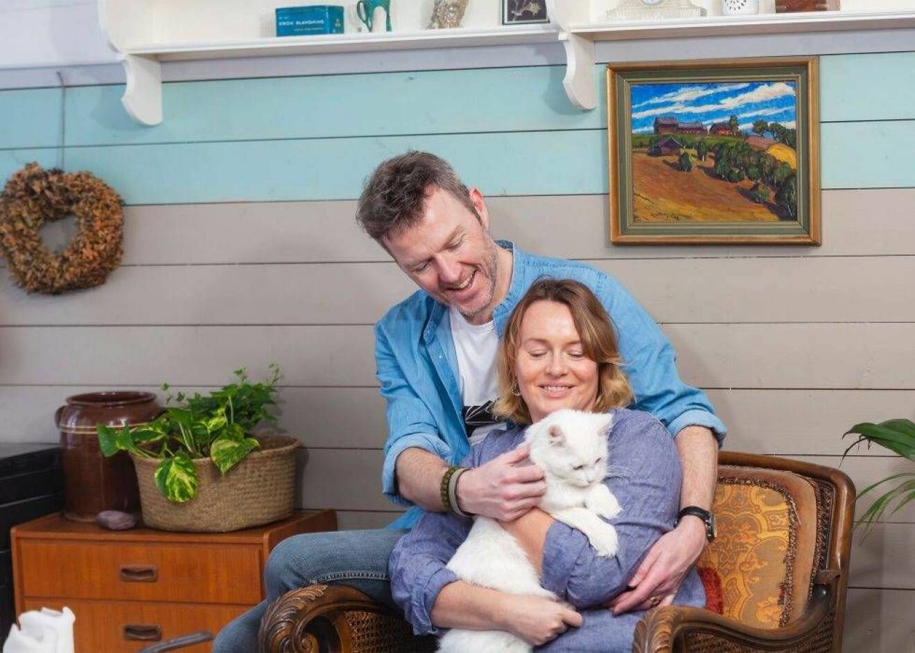 En kvinna i 45-årsåldern sitter med en katt i knät och hennes make bakom sig.