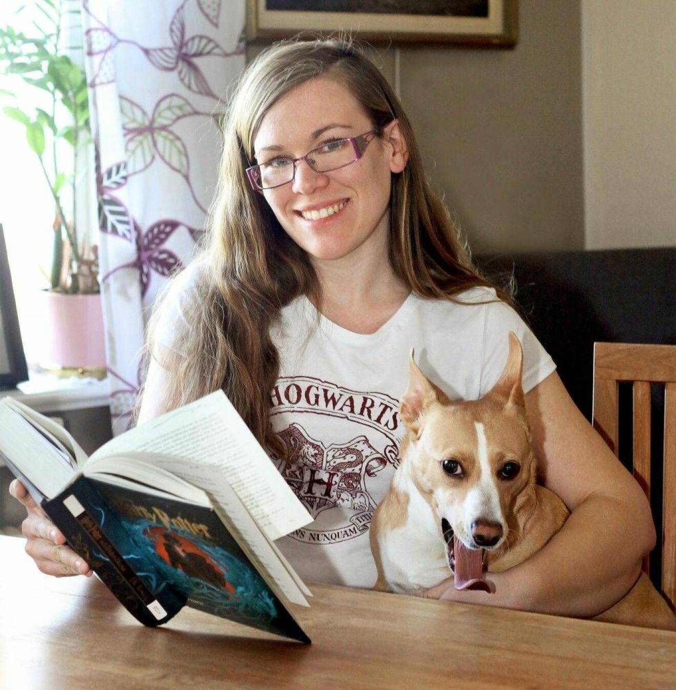En ung kvinna sitter vid ett bord med sin hund och läser Harry Potter-bok.