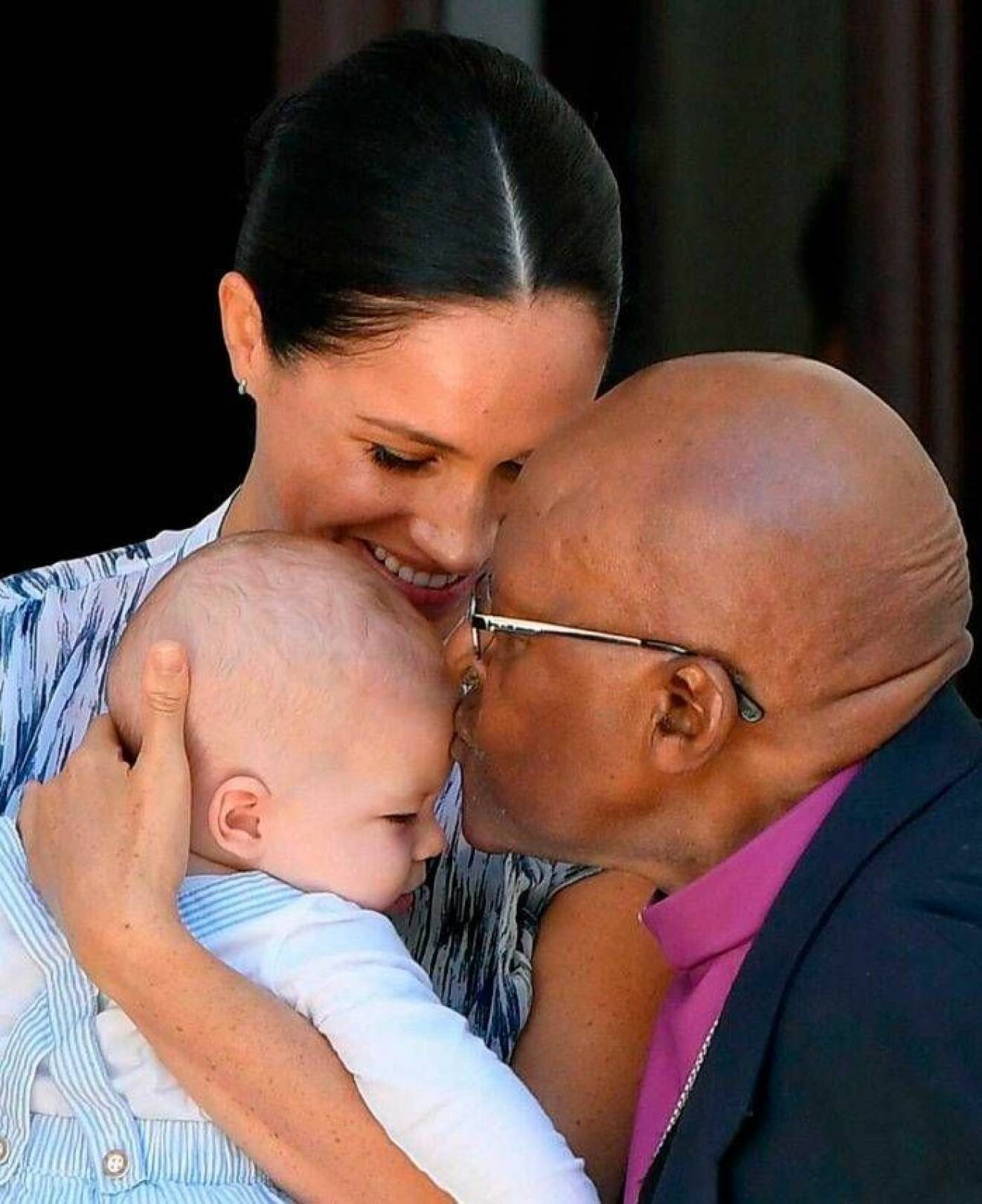 Desmond Tutu pussar Archie på pannan medan mamma Meghan nöjt ser på.