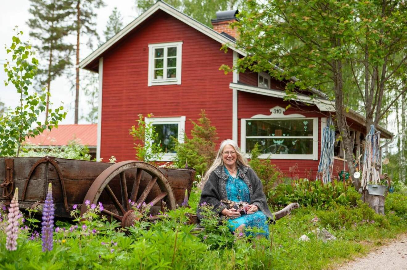 Sacha, en äldre kvinna i långt grått hår, sitter i grönskan framför ett rött hus.