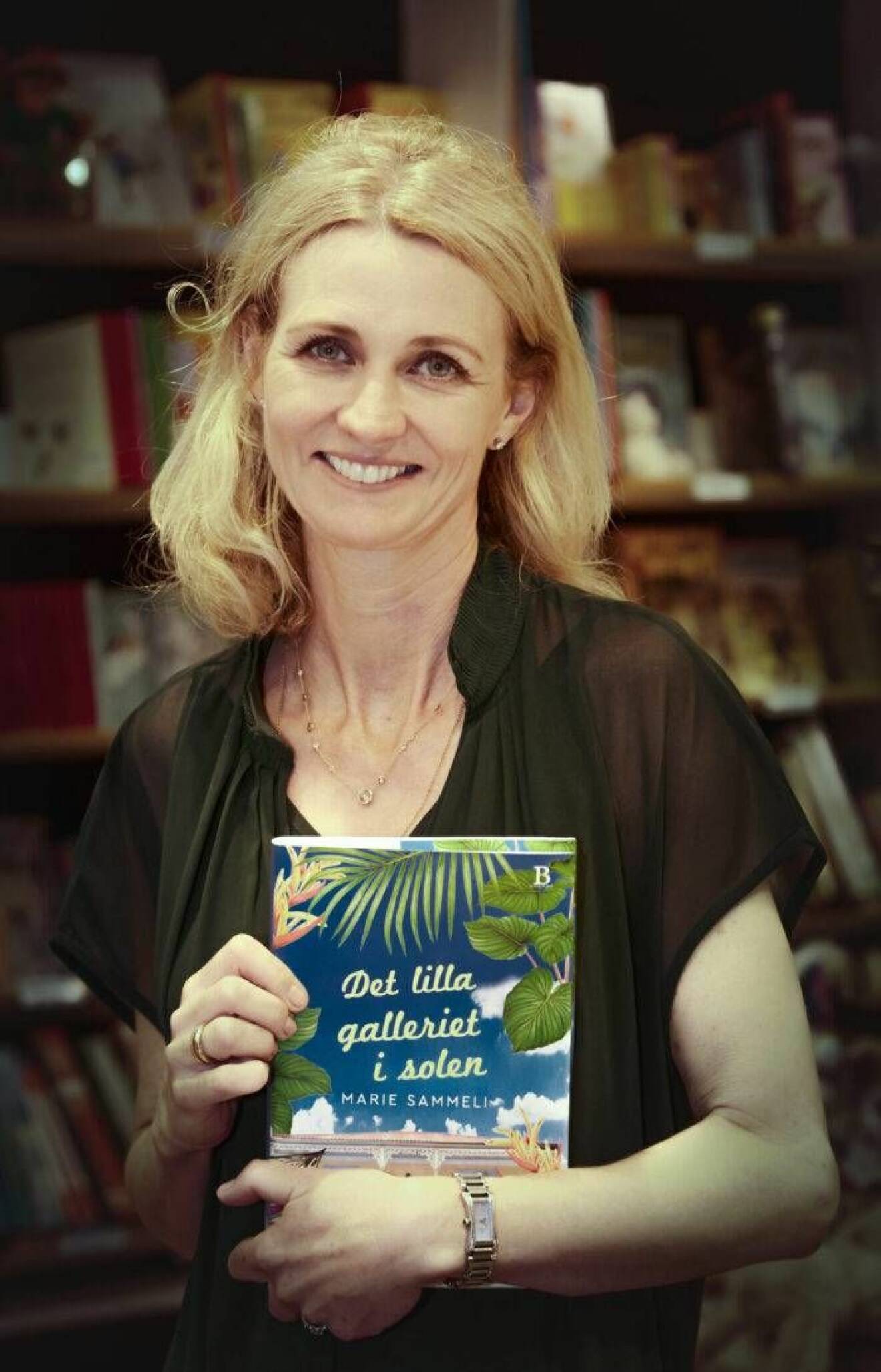Marie Sammeli står i en bokhandel med sin egen bok Det lilla galleriet i solen i famnen. 