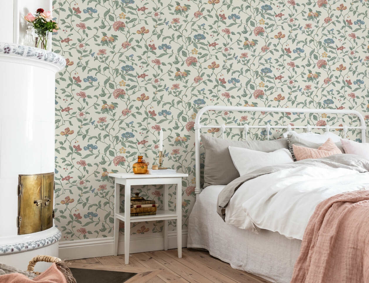 Ett sovrum med en vit kakelugn, brädgolv och en säng med fluffiga ljusa lakan. Väggarna är täckta i en krämfärgad tapet med slingrande blomsterrankor.