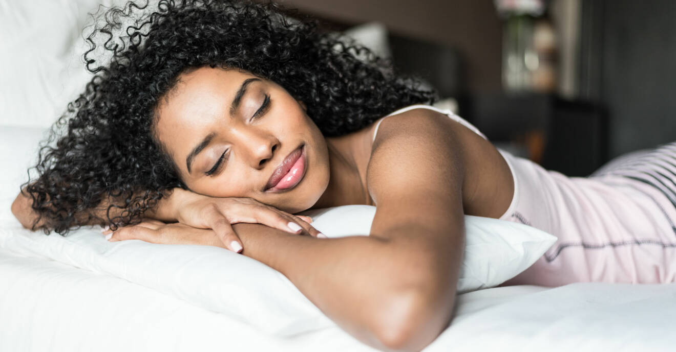 En kvinna ligger på en säng och sover med händerna under huvudet.