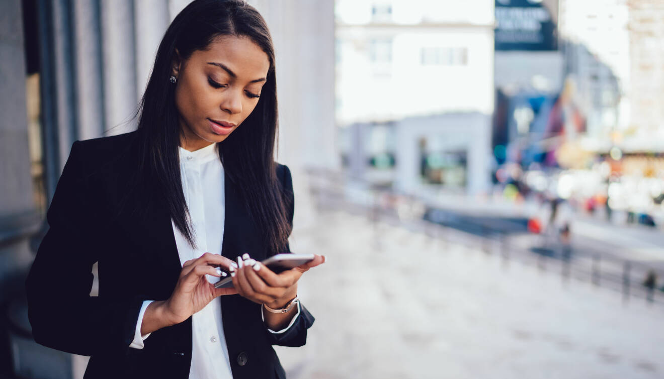 En afroamerikansk kvinna i kostym ser allvarligt ner i sin mobiltelefon.