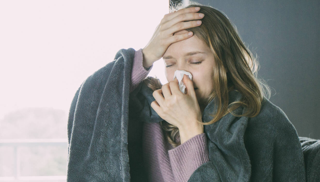 Kvinna med förkylningssymtom som snyter sig och håller sig för pannan.