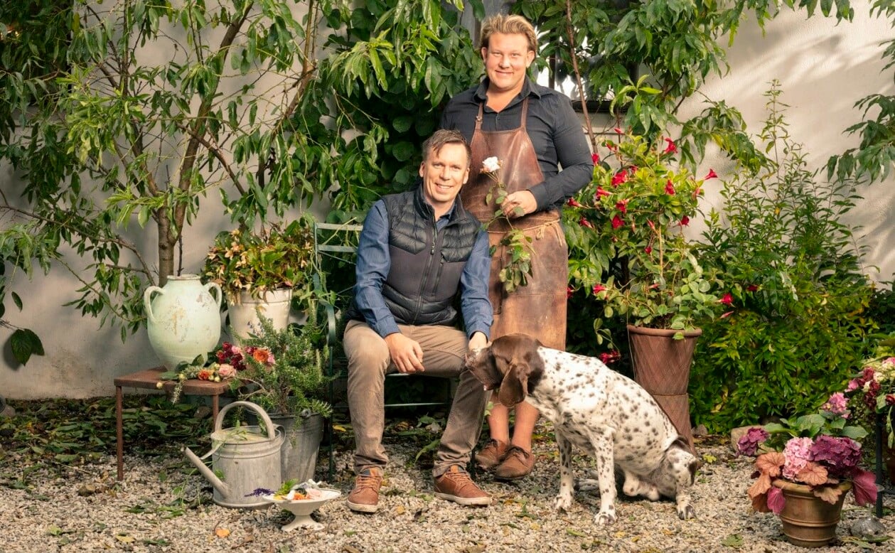 Tre av fem i familjen på Eklaholm: Karl Fredrik Gustafsson, sambon Petter Kjellén och hunden Prins Hamlet. Foto: Foto: Jakob Dahlström/TV4. Karl Fredrik med en vit ros, sambon Petter och hunden i en grön och prunkande miljö.