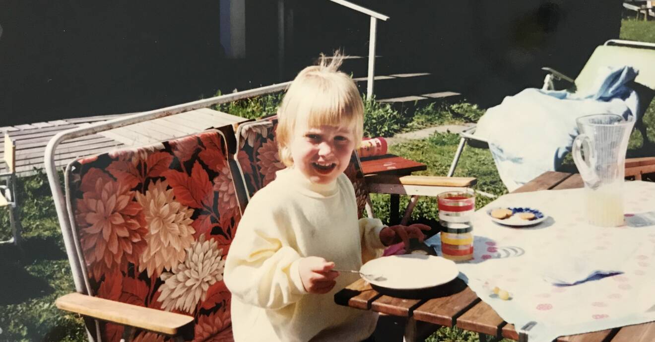 Lilla Åsa sitter vid ett trädgårdsbord och fikar.
