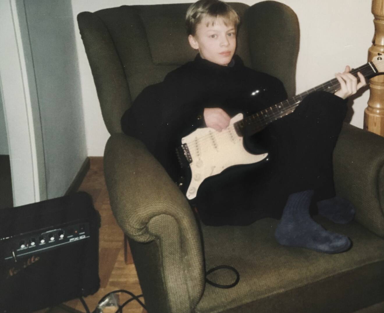 Björn Dixgård var i tolvårsåldern när han lärde sig att spela gitarr och här sitter han uppkrupen i grönbrun en öronlappsfåtölj av äldre snitt med en elgitarr i knät och på golvet bredvid står en förstärkare.